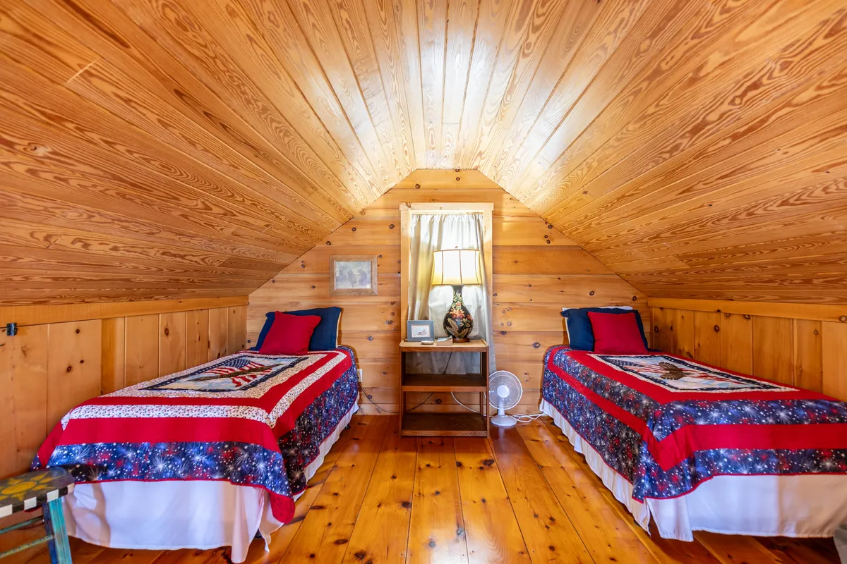A whimsical loft space with two twin beds adorned with patterned quilts, a wooden ceiling, and a bedside table with a lamp. The setting is warm and playful, ideal for kids or friends, with a cozy ambiance for reading or sharing bedtime stories.