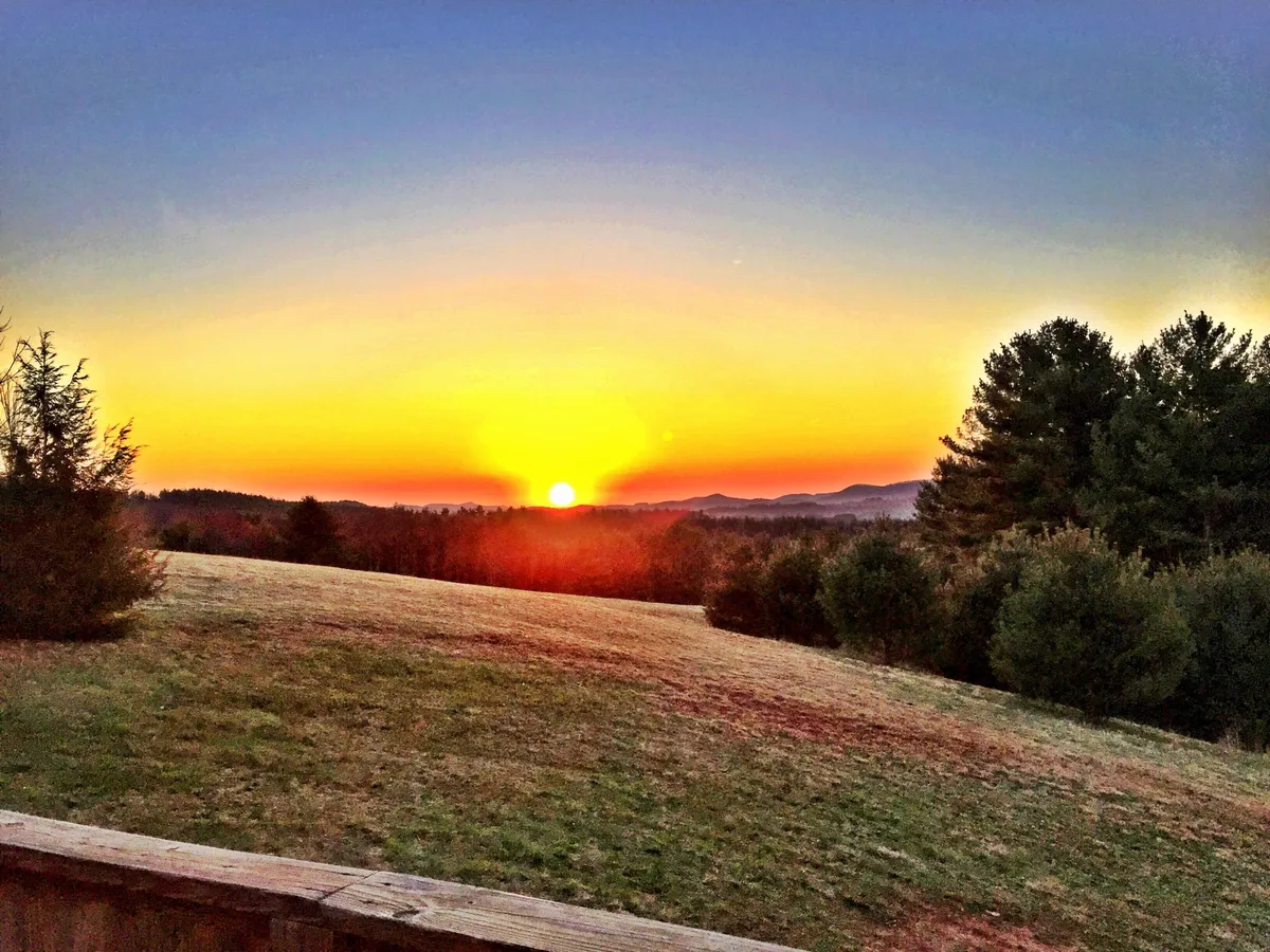 A breathtaking view of a vibrant sunset over distant mountains and a tree-lined horizon. The sky is painted in warm hues, creating a tranquil backdrop ideal for peaceful moments and evening reflection. The scene invites viewers to imagine watching sunsets from the deck or porch.