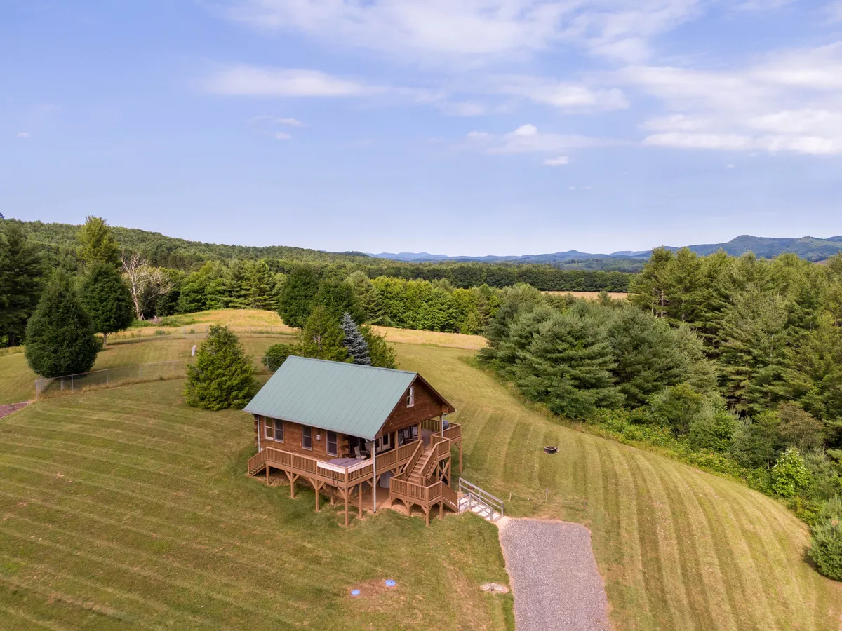 This image showcases the wooden cabin with its green metal roof nestled amid lush, wooded surroundings. The surrounding landscape includes open lawn space and nearby woods, evoking the sense of exploring nature right from the doorstep.
