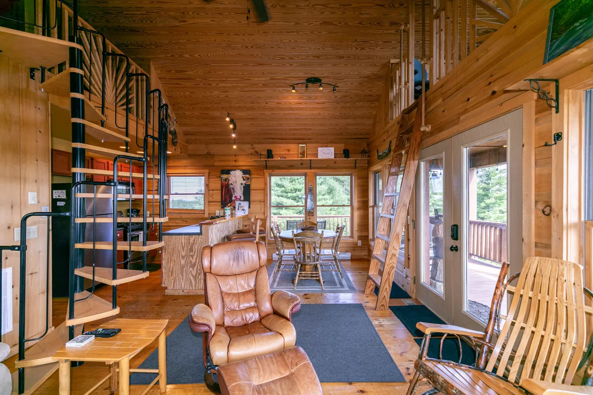 A cozy interior featuring a spiral staircase, a spacious wooden dining table, a comfortable armchair, and large windows offering scenic views. The open layout suggests a seamless connection between dining and relaxation, with plenty of space for family gatherings and shared meals.