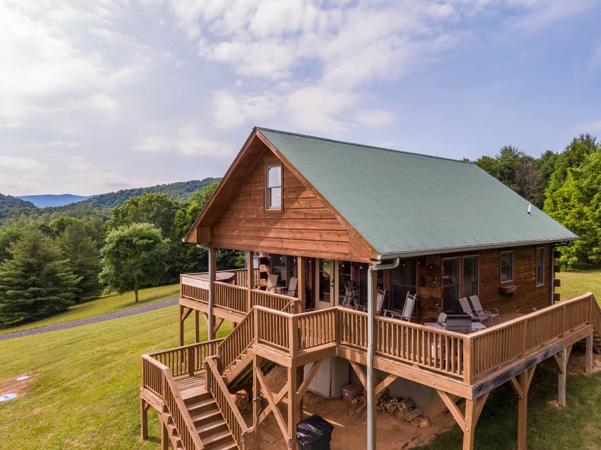 The image showcases a charming wooden cabin with a green roof perched atop a gentle knoll, surrounded by lush natural landscape. The expansive wraparound deck and stairs invite guests to relax, while the covered porch provides the perfect spot to soak in sweeping sunset views. The tranquil setting hints at the privacy and connection to nature that the property offers.
