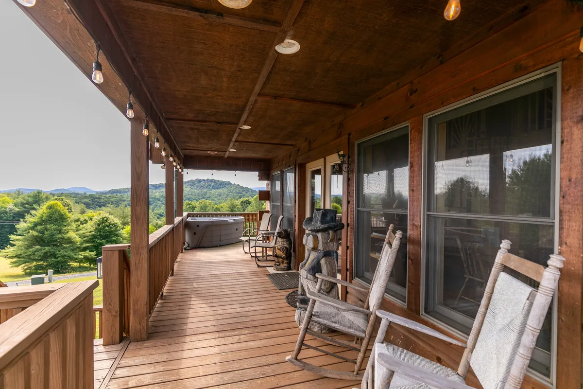 A cozy porch scene featuring classic wooden rocking chairs and a bubbling hot tub, all set against a breathtaking mountain view. String lights hang above, and the golden hues of sunset reflect on the mountains beyond. The porch’s wooden railing and deck add rustic charm, creating a space that feels both private and intimately connected to nature.