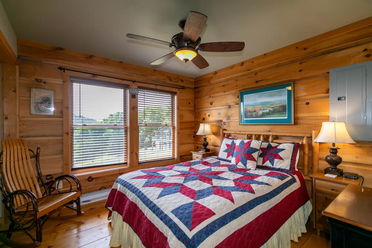 A cozy master bedroom featuring a king-size bed with a quilted bedspread, wooden walls, windows with blinds that let in natural light, a rocking chair, and table lamps on either side of the bed. The room feels warm, restful, and inviting, with sunlight streaming in to offer a gentle start to the day.