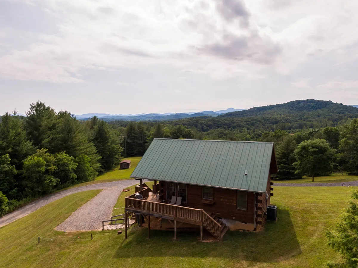 An inviting log cabin with a distinctive green metal roof, surrounded by lush trees and a panoramic mountain backdrop. The covered porch is visible, with outdoor seating that promises tranquil mornings or evenings. The scene is bathed in soft, natural light, and the setting is peaceful, hinting at sunrise views and wildlife encounters.