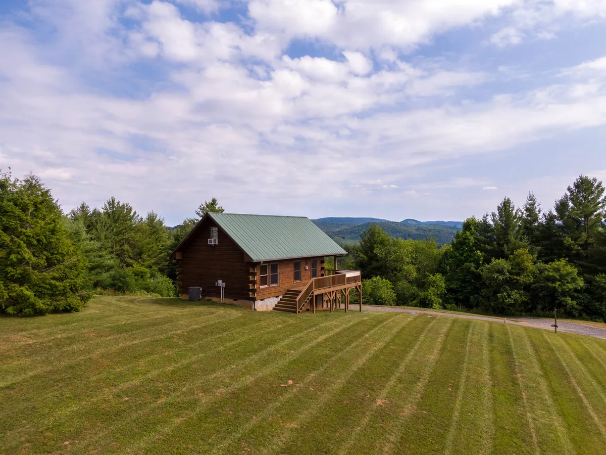 A serene exterior shot capturing the log cabin with its green metal roof, expansive lawn, and distant mountains. The scene exudes tranquility and highlights the cabin’s spacious outdoor area, perfect for gatherings, play, and soaking in panoramic views. The wraparound deck and covered porch are visible, framed by lush trees and set against a stunning mountain backdrop.
