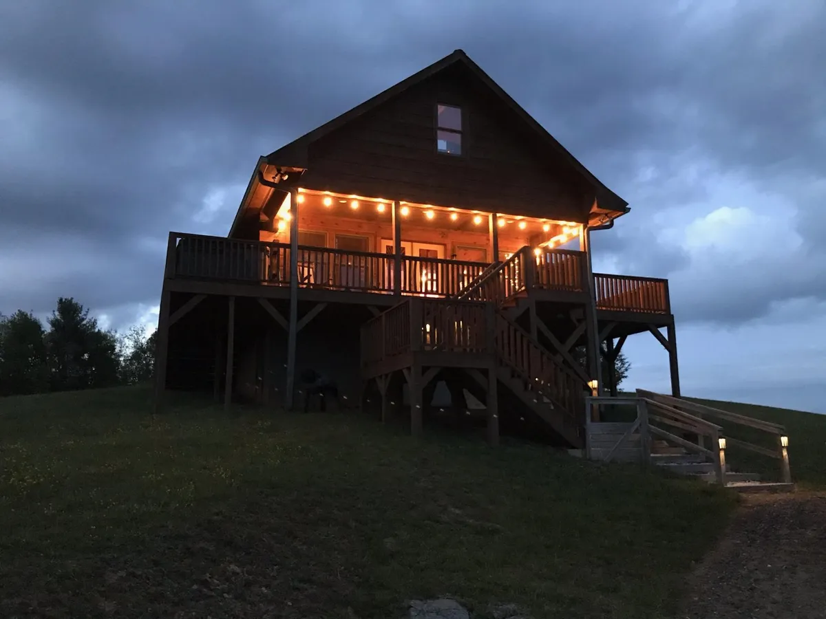 A warmly illuminated cabin porch with a clear view of the surrounding landscape, inviting guests to relax outside during the evening. The setting hints at stargazing and quiet moments after a day of exploring with pets.