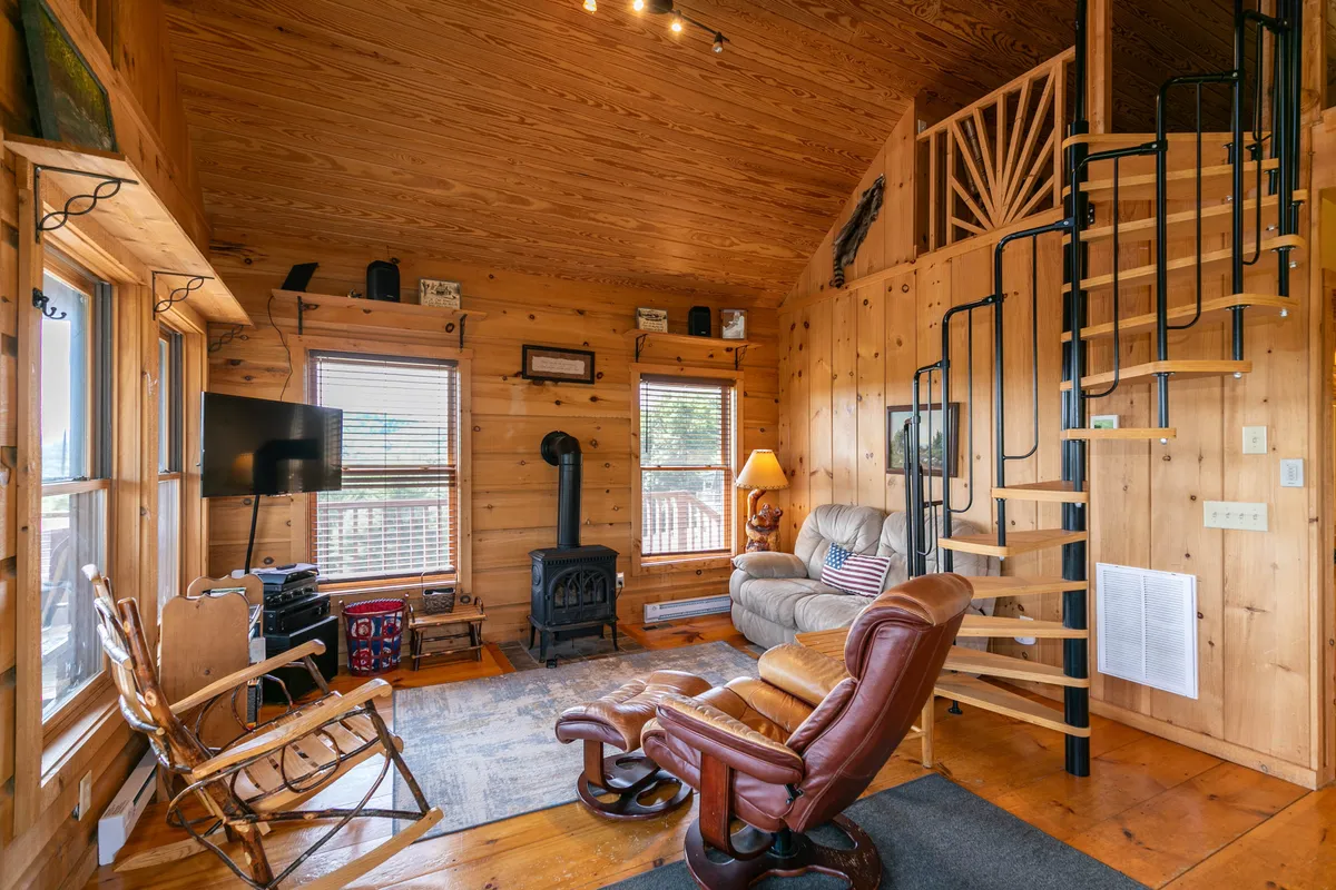 This image showcases the heart of the cabin's great room, featuring inviting wooden walls, a spiral staircase, a cushy armchair, a flat-screen television, and a wood-burning stove. Natural light filters through the windows, highlighting the warm tones of the hardwood floors and rustic charm.