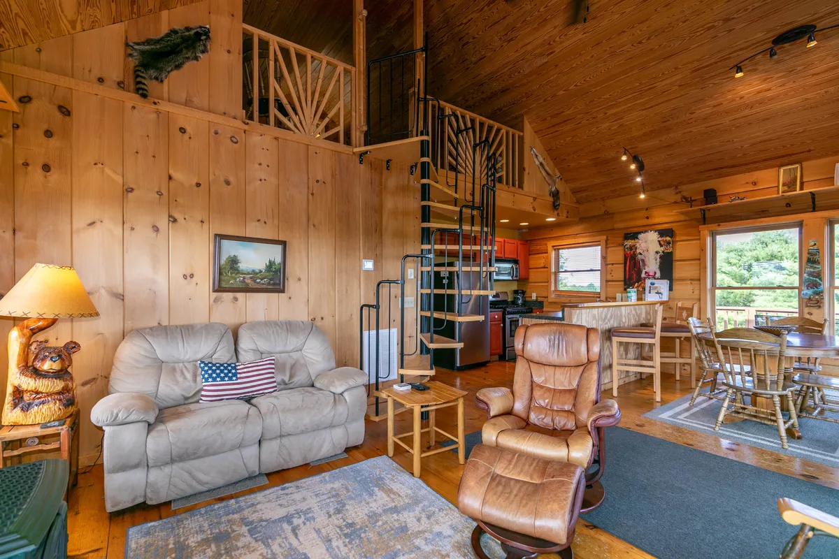 The view offers a different angle of the open-concept great room, highlighting a leather sofa, spiral staircase, and wooden dining table with a decorative lamp, all set against warm wooden walls. Sunlight streams in, reinforcing the room’s bright and welcoming feel.
